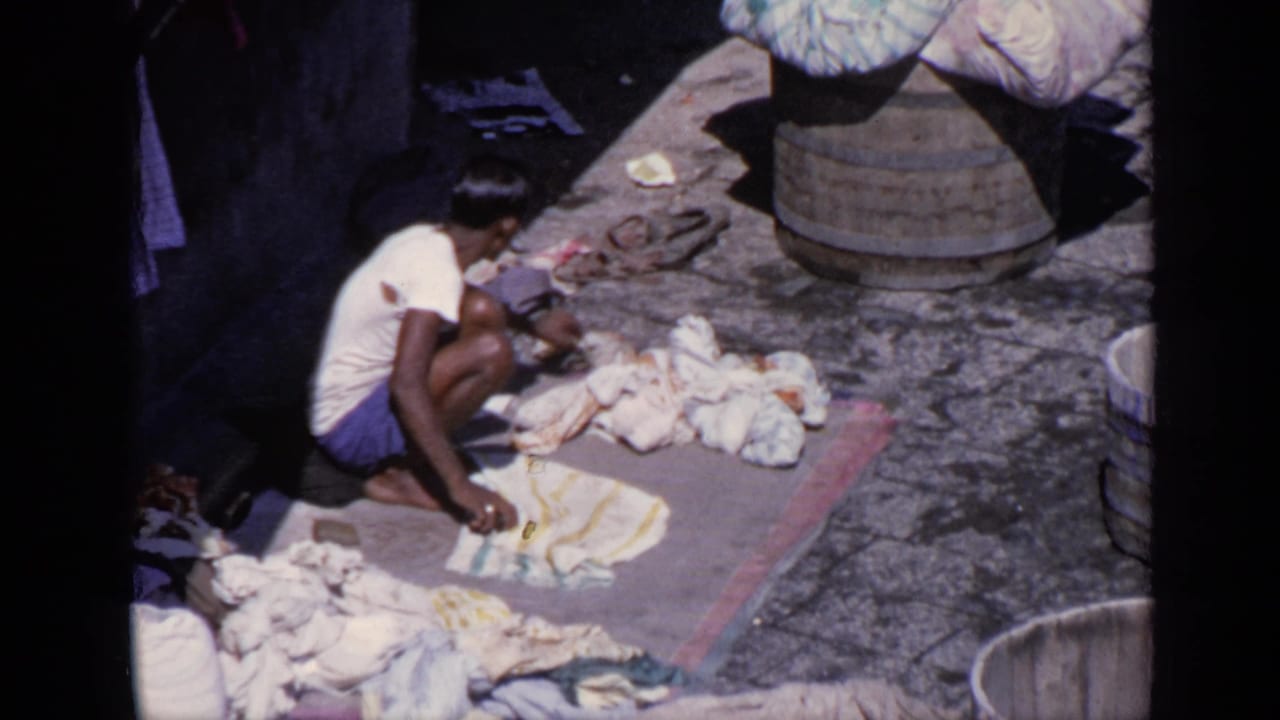 Man crouches on a mat washing clothes beside piles of laundry and wooden barrels.