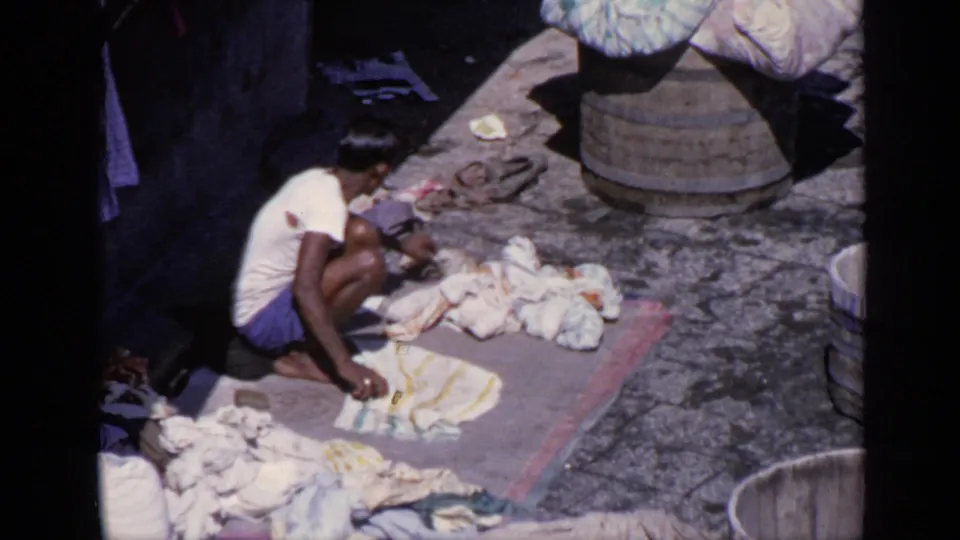 In 1970 India home-movie footage, a man crouches on a mat scrubbing garments in a stone courtyard near laundry piles and wooden barrels.