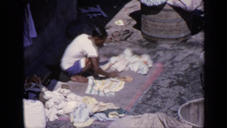 A man in 1970 India continues scrubbing cloth on a mat, with pale garments, stone paving, and large wooden tubs around the work area.