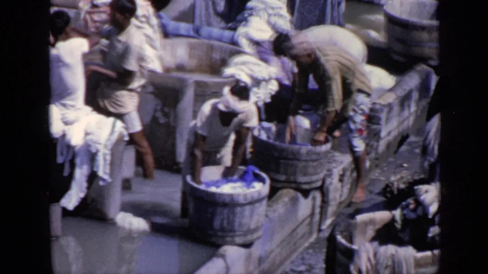 Several men work at a communal washing area in India, using large wooden tubs and a stone basin while garments hang and pile nearby.