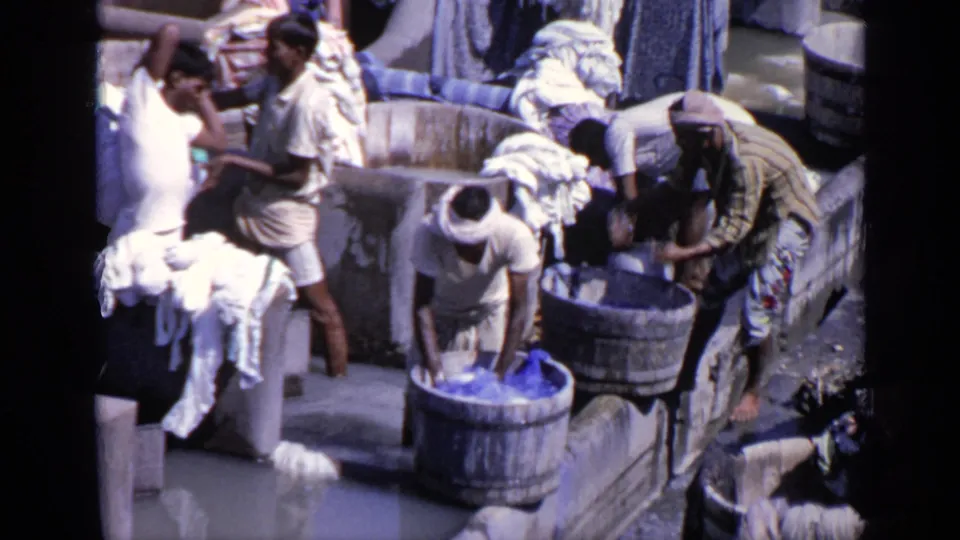 Men at a communal laundry space in India handle wet garments around wooden tubs, a stone basin, hanging cloth, and piles of washed fabric.