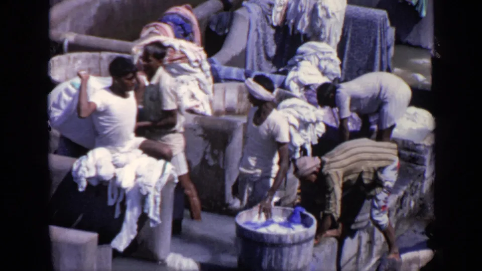 Men continue hand-washing and handling laundry in an outdoor wash area, with textiles piled and draped around the basins.
