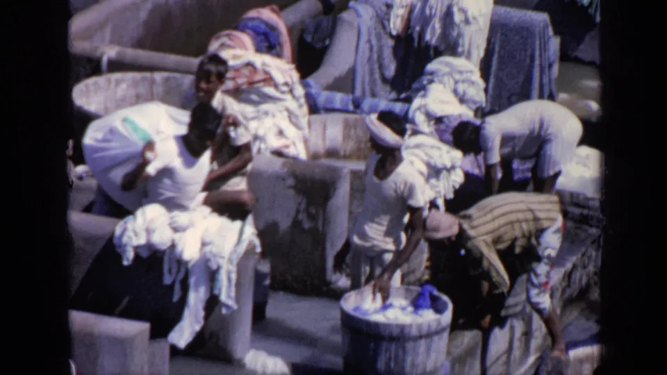 1970 India home-movie frame of men washing and sorting clothes among crowded tubs, basins, and draped laundry.