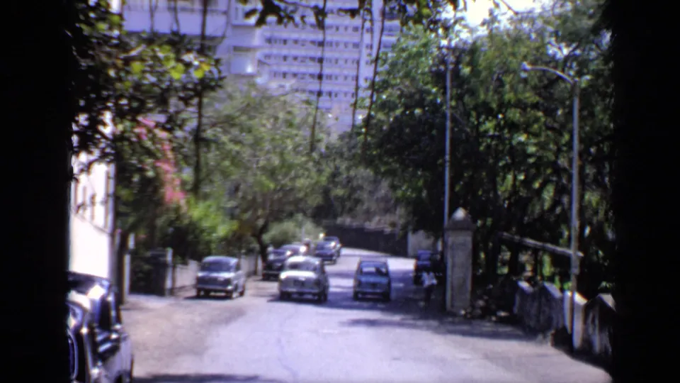 1970 India 8mm street view with trees, parked cars, low walls, and a large building visible beyond the road.