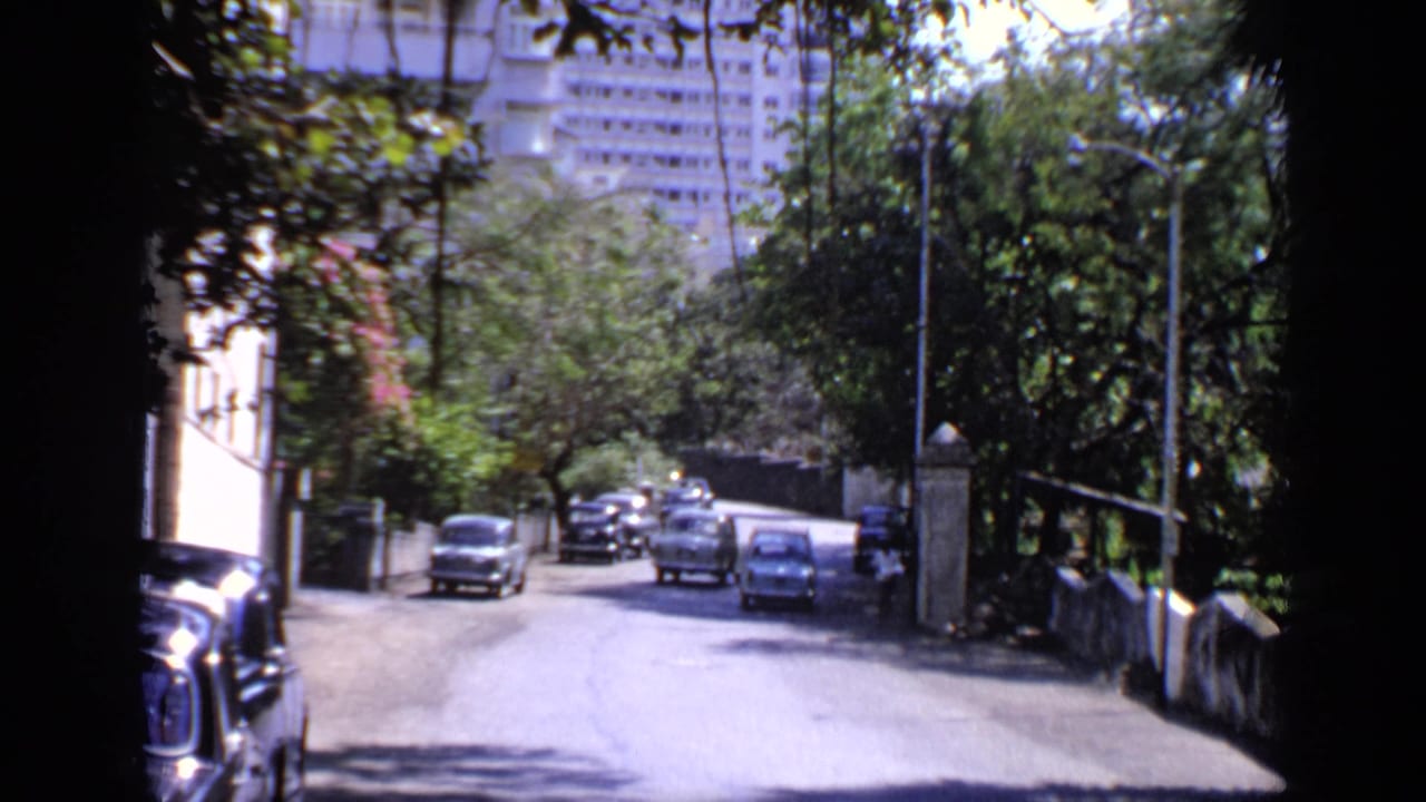 1970 India 8mm frame showing cars on a narrow tree-lined street with a pale apartment block in the background.
