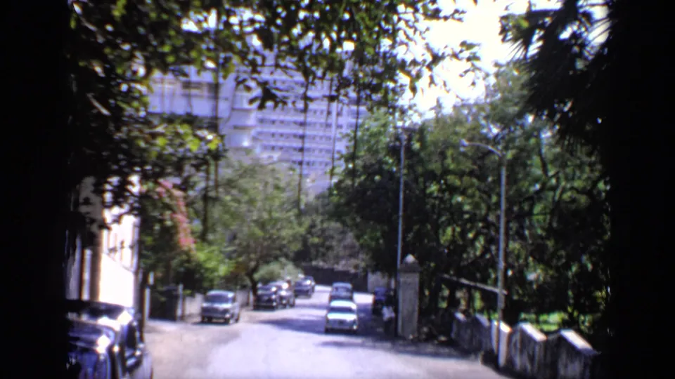 1970 India 8mm frame of cars moving on a sunlit road framed by trees, walls, and distant apartment buildings.
