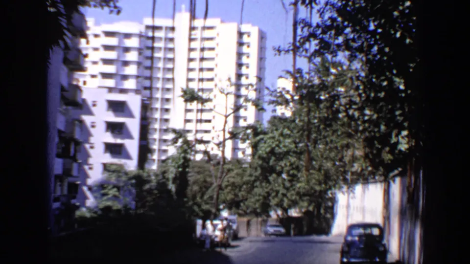 1970 India 8mm frame showing a white apartment block, roadside trees, pedestrians, and a dark car on a narrow street.
