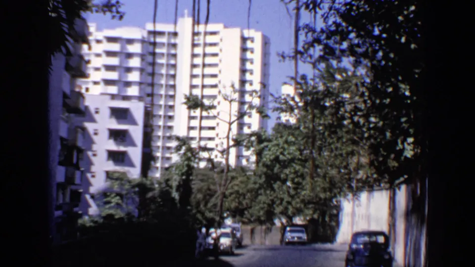 1970 India 8mm frame with a blue car near a roadside wall, trees, pedestrians, and a white apartment building.