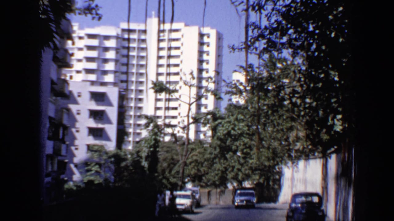 1970 India 8mm frame showing a shaded street, parked cars, trees, and high-rise apartment or hotel buildings beyond.