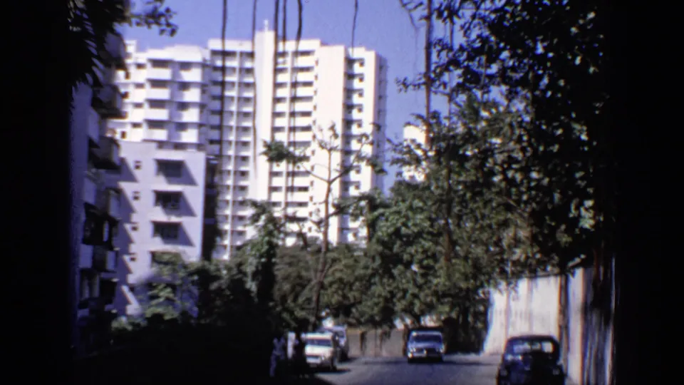 1970 India 8mm frame showing a shaded street, parked cars, trees, and high-rise apartment or hotel buildings beyond.