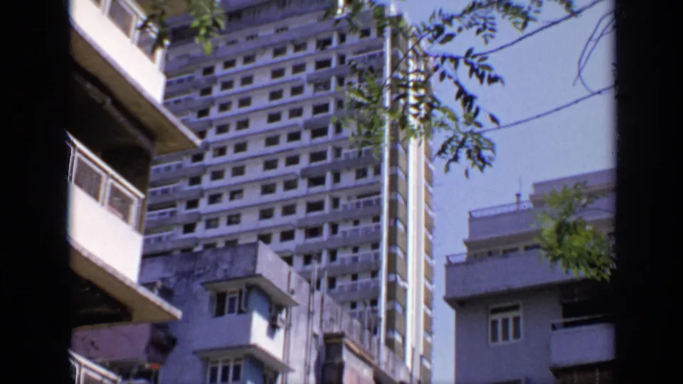 1970 India home-movie frame showing a tall high-rise, neighboring apartment balconies, tree branches, and blue sky.