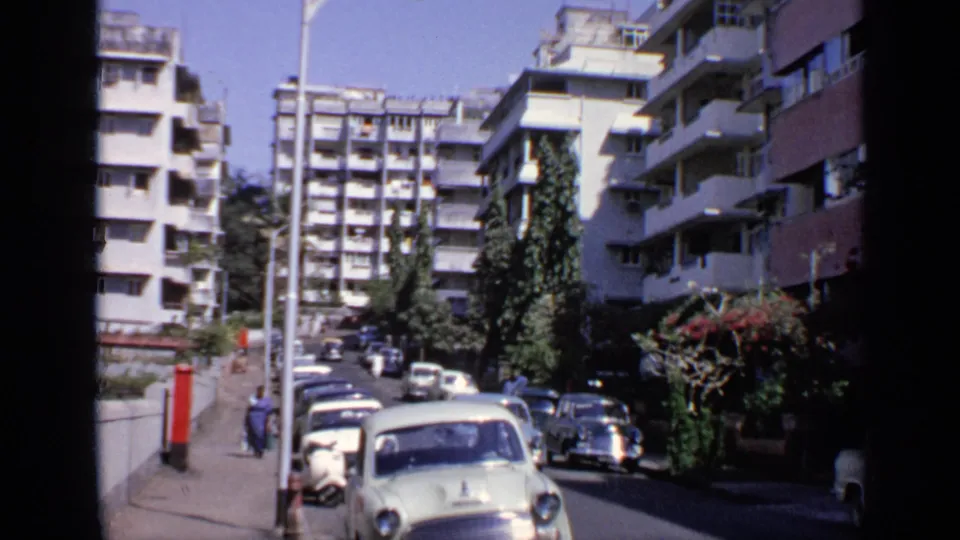 1970 India street scene with a light blue classic car, parked vehicles, pedestrians, trees, and high-rise apartment buildings.