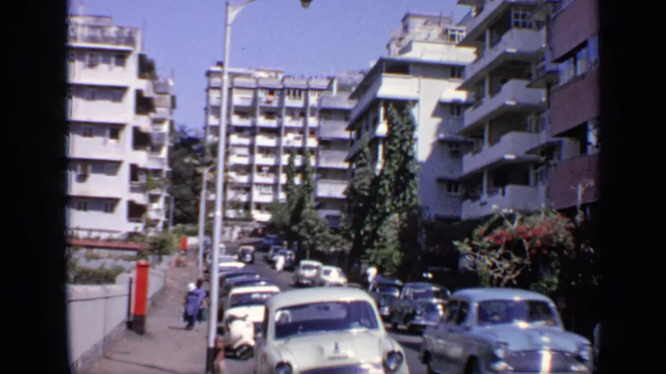 1970 India 8mm view of classic cars on a residential street bordered by tall apartment buildings, trees, and pedestrians.
