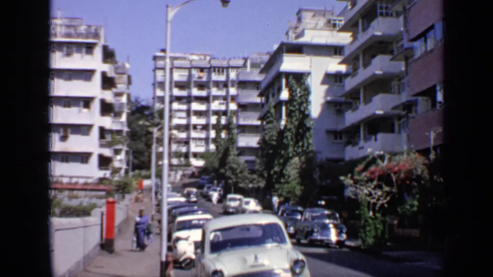 1970 India street view with a light blue car passing parked vehicles under tall white apartment buildings and greenery.