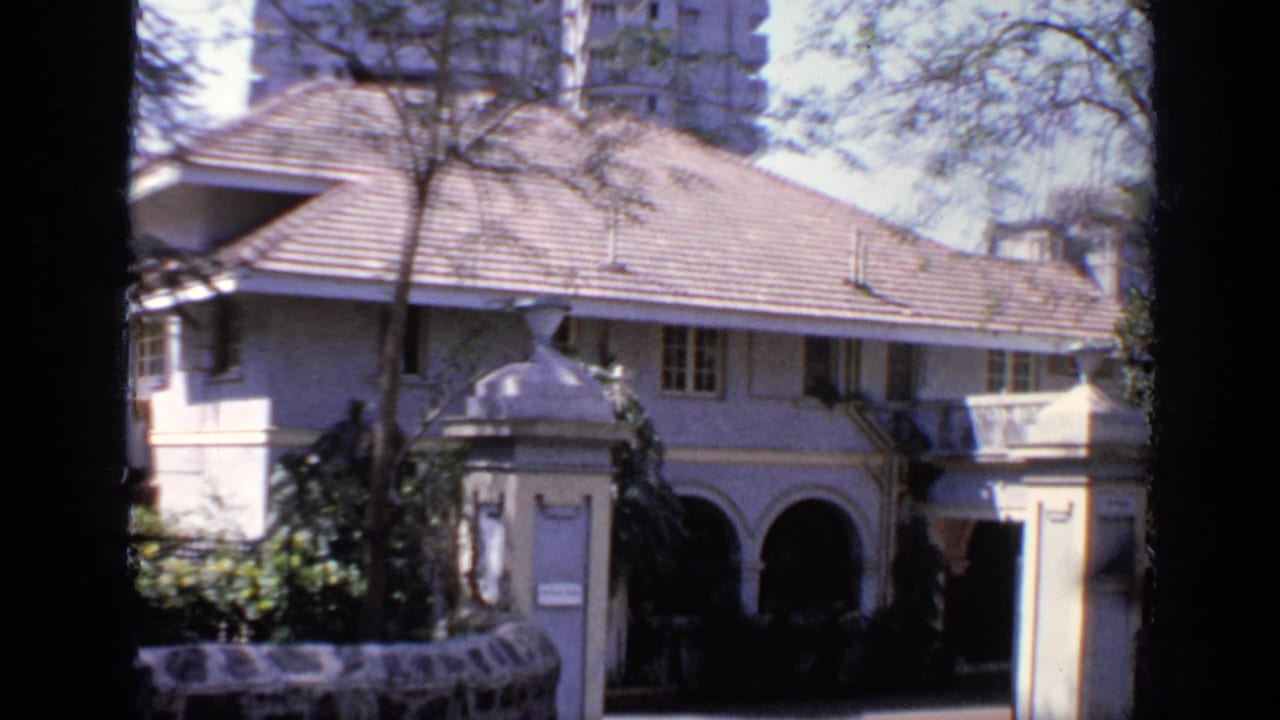 White house with tiled roof, arched entry, trees, and tall buildings behind it.