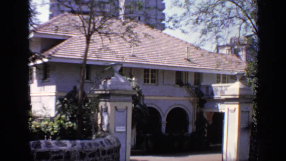 1970 India 8mm frame with the same pale house exterior, tiled roof, arched entrance, surrounding trees, and high-rises behind.