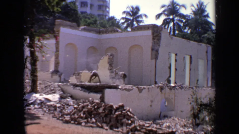 1970 India 8mm frame showing a partially demolished pale building or wall, rubble piles, workers, palms, and urban buildings beyond.