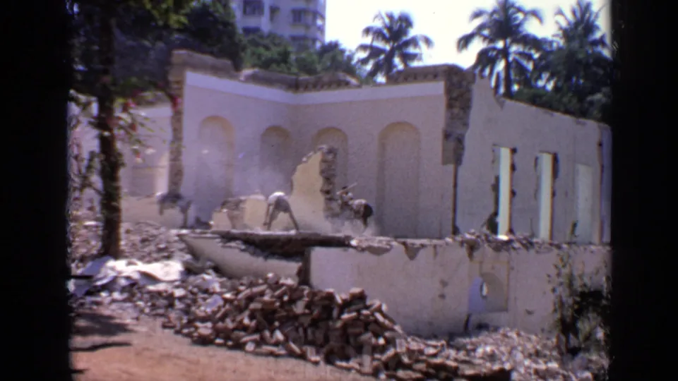 1970 India 8mm frame of workers near rubble and a broken pale wall, with palm trees, bright sunlight, and nearby urban buildings.