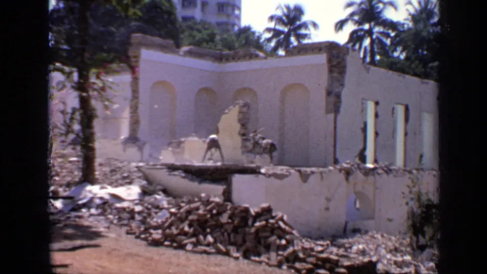 1970 India 8mm frame with a damaged wall, rubble on the ground, tropical trees, and a worker visible near the broken structure.