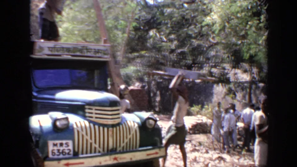1970 India 8mm frame showing a blue truck with a Hindi roof sign as men load long wooden planks in a shaded roadside area.