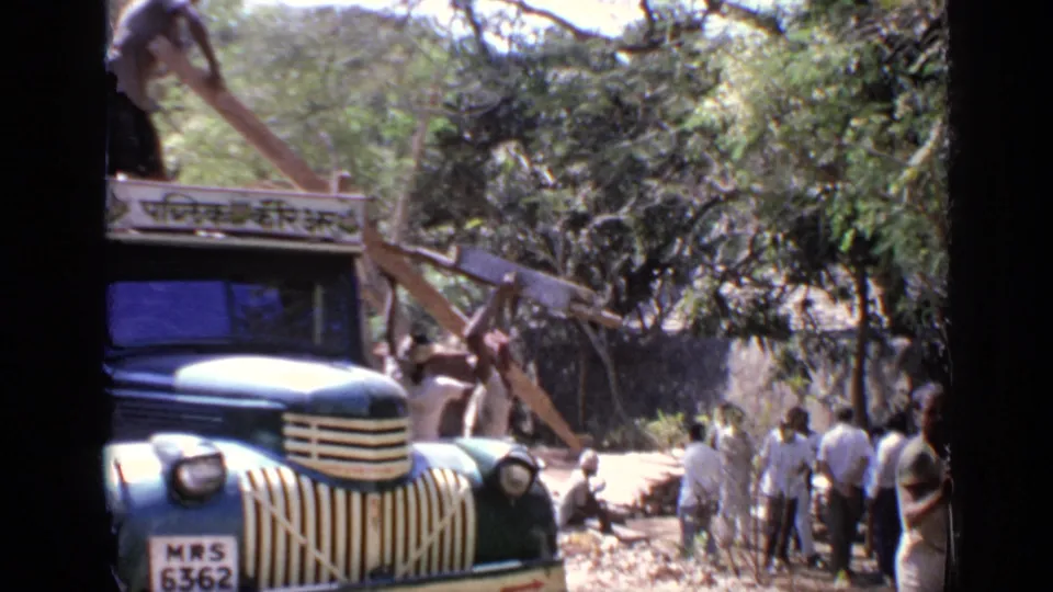 1970 India 8mm frame of men handling long wooden planks around a blue commercial truck, with trees and a wall in the background.