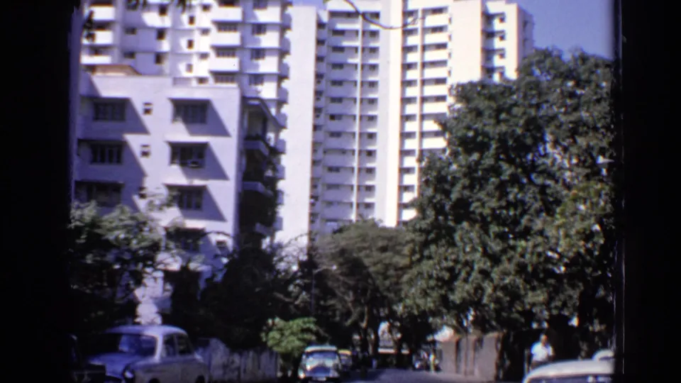 1970 India street view with white apartment blocks, leafy trees, parked cars, and distant pedestrians in soft 8mm focus.