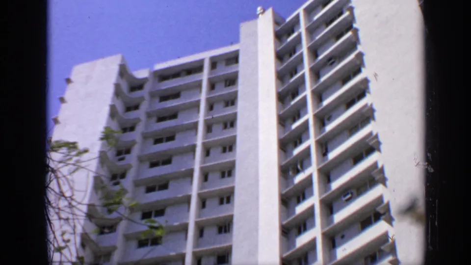 1970 India home-movie frame looking upward at a white high-rise apartment building with balconies and small tree branches.