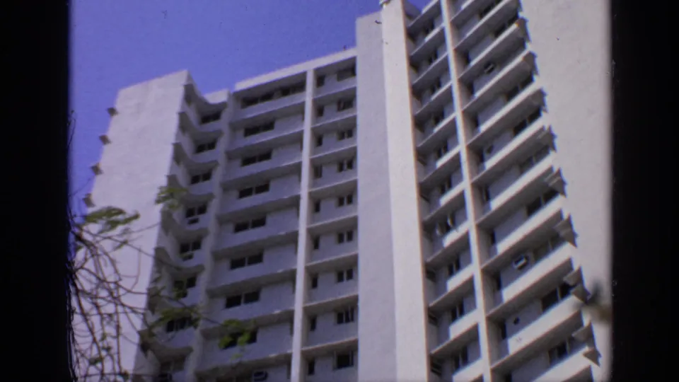 1970 India 8mm frame of a white apartment tower seen from below, with balcony rows, shadowed windows, and sparse branches.
