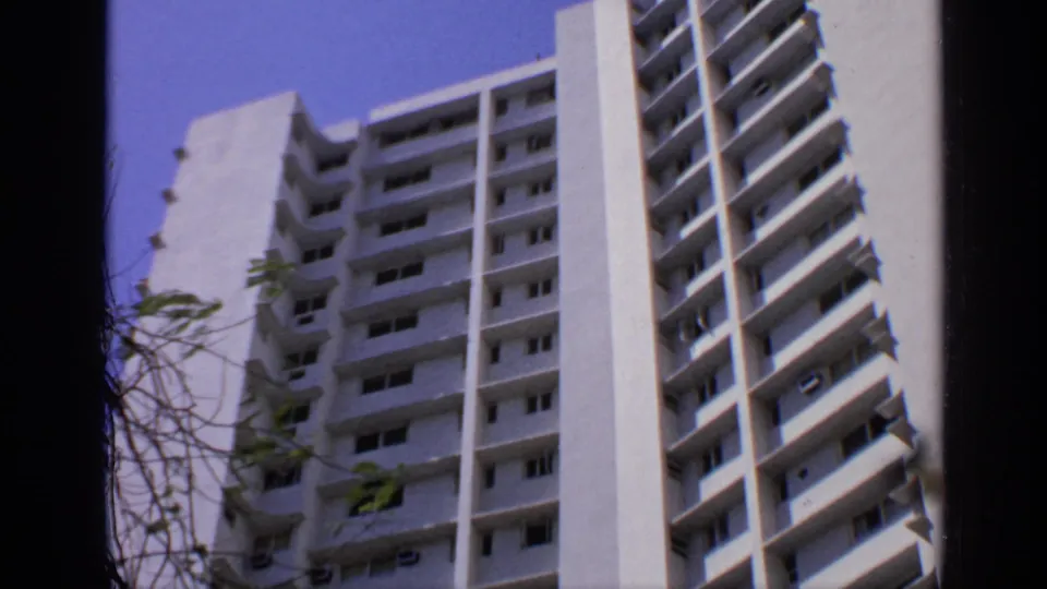 1970 India home-movie frame showing a white high-rise apartment building from below, with balconies and a clear blue sky.