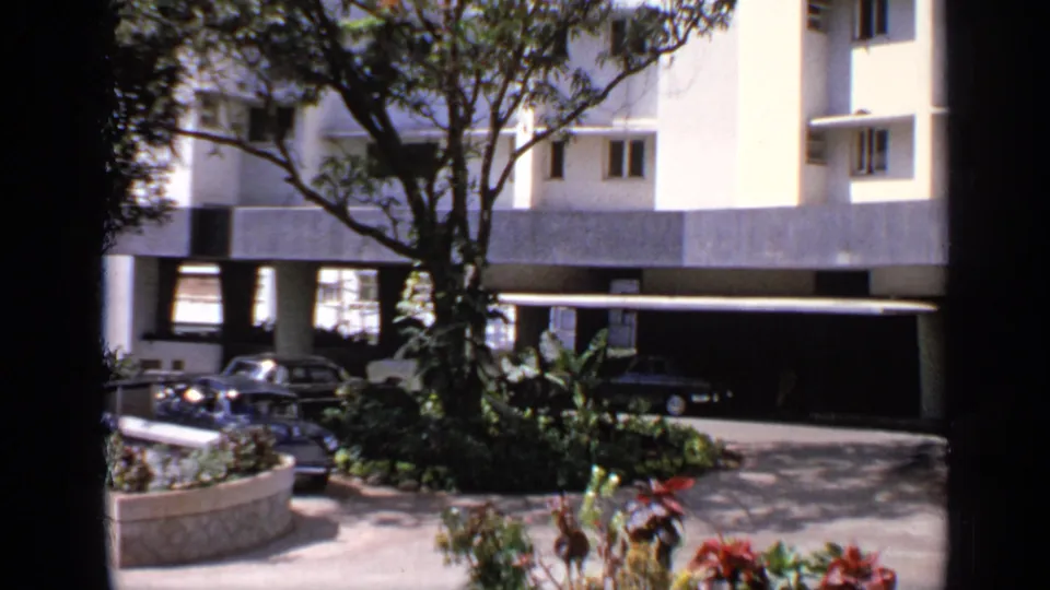 1970 India home-movie frame showing a shaded residential or hotel courtyard with parked cars, greenery, and a covered entrance.
