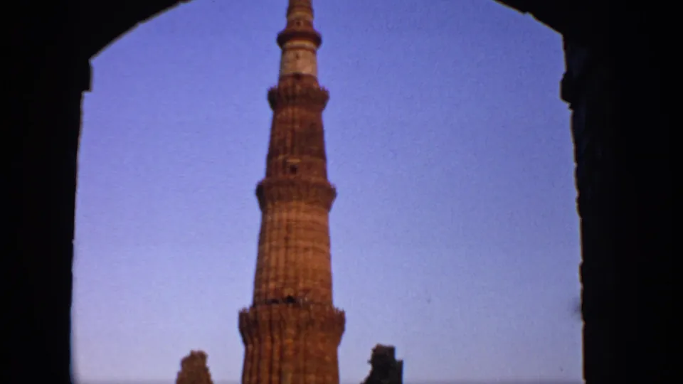 1970 India home-movie frame showing the Qutub Minar rising through a shadowed archway, with the tower silhouetted against a blue-purple sky.