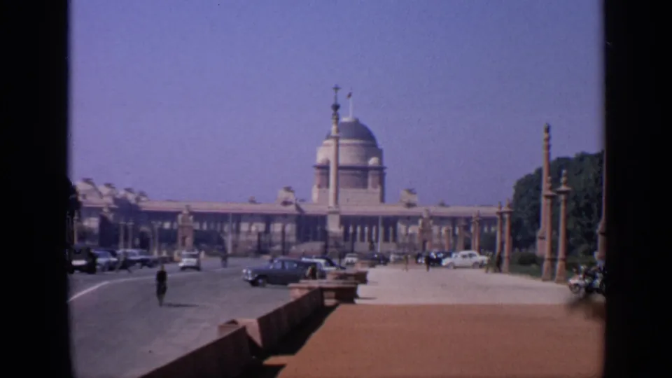 Wide 1970 India street view of a domed building, likely Rashtrapati Bhavan, with classic cars and small pedestrians on the boulevard.