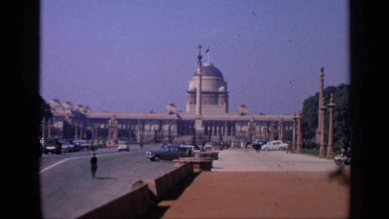 1970 India home-movie frame showing a domed civic-style building across a broad road with cars, pedestrians, pillars, and trees.