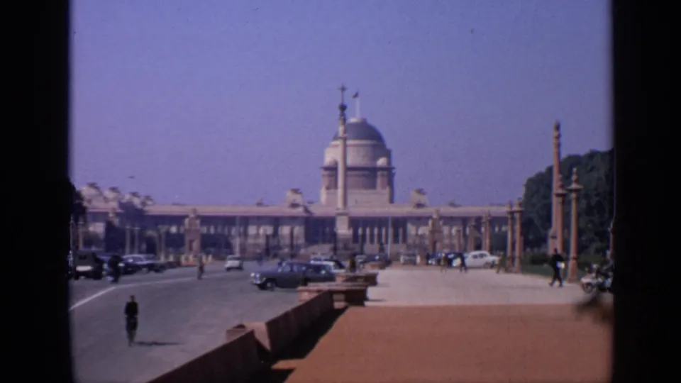 1970 India street view with a domed building, cars crossing the road, small pedestrians, roadside pillars, and trees at the edge.
