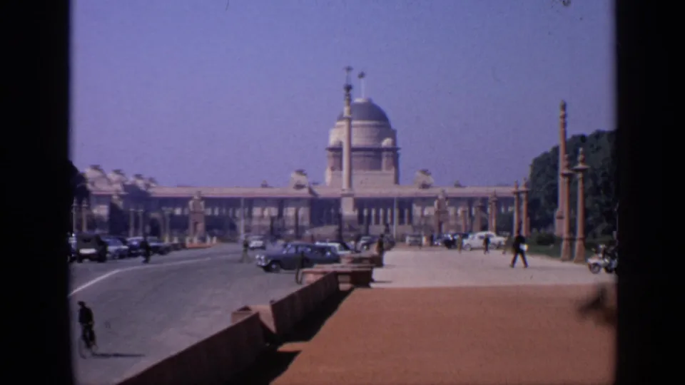 1970 India frame of a formal domed building viewed across a wide roadway with cars, a cyclist, pedestrians, pillars, and trees.