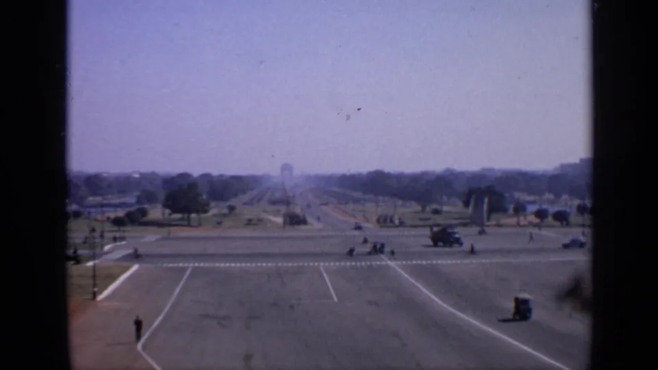 1970 India home-movie frame looking down a broad formal avenue with distant vehicles, pedestrians, trees, and open paved lanes.