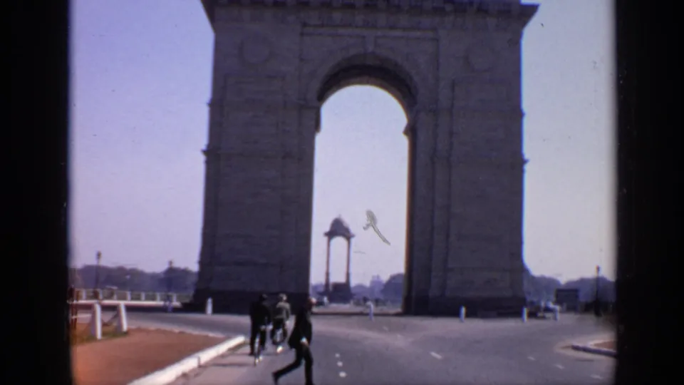 1970 India home-movie frame showing India Gate beyond a wide road as several pedestrians move near the foreground.