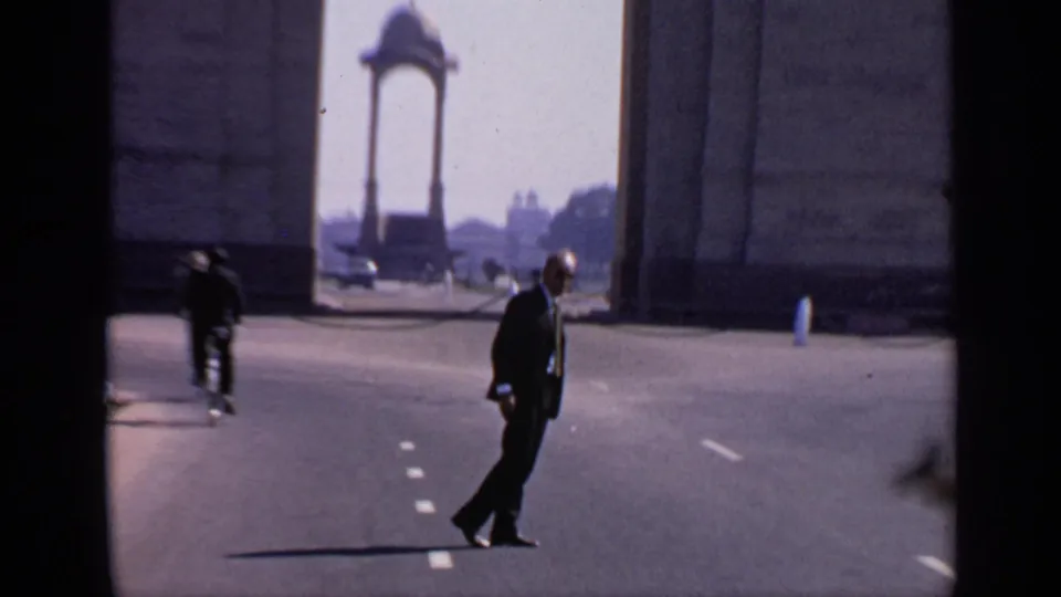 A suited man walks across a broad road near India Gate, with another figure and distant traffic visible in the background.