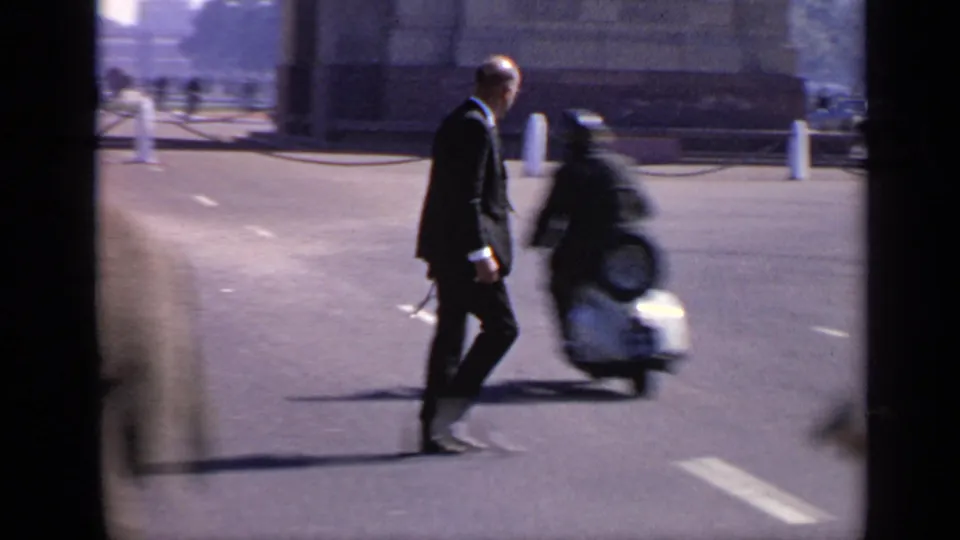 The suited pedestrian continues across the roadway while a scooter passes close by in the foreground near India Gate.