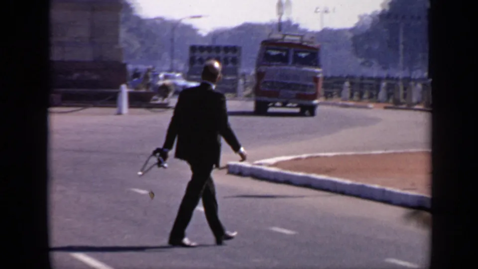 The man in a dark suit walks across the open road as a red bus and other traffic appear near India Gate in the background.