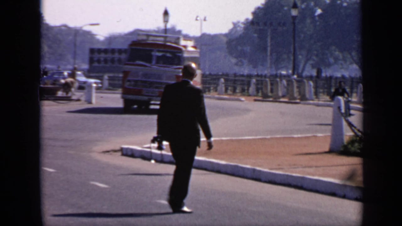 Man in a dark suit crossing a sunlit road as a red and white bus approaches in the background.