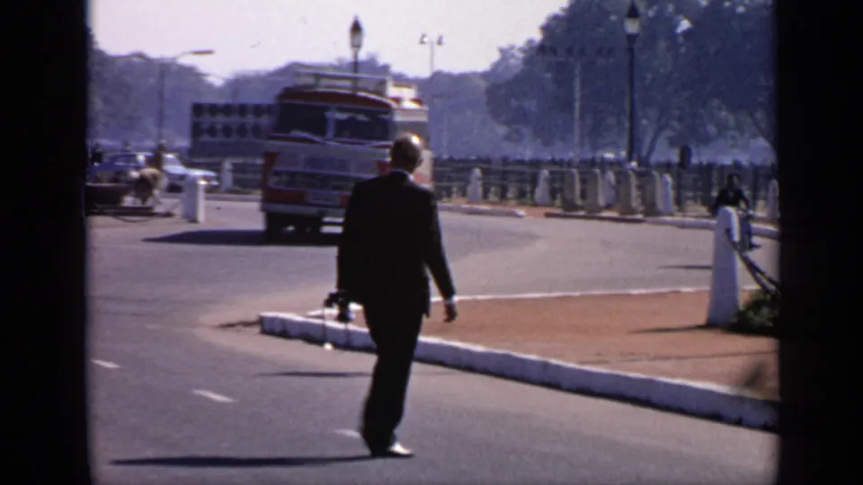 1970 India street view with a suited man crossing the road, a red and white bus, white curb, lampposts, and distant trees.