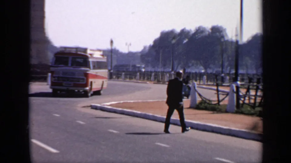 1970 India roadway scene with a man crossing near roadside posts and railings while a red and white bus travels behind him.