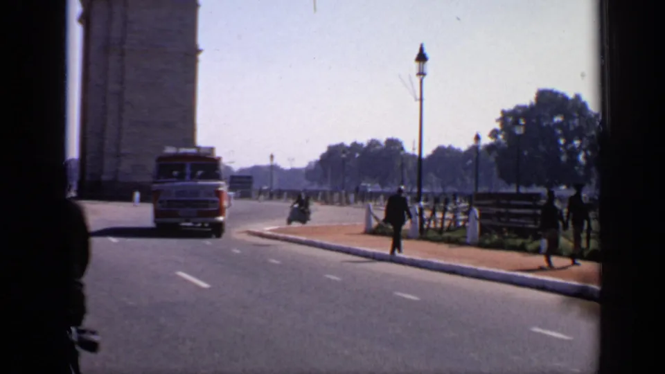 Delhi landmark approach in 1970 with a bus near the base of India Gate, pedestrians, roadside railings, lampposts, and open lanes.