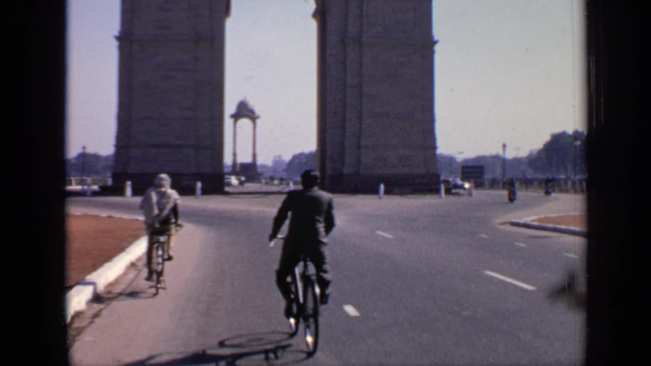 Two cyclists travel on the broad road near India Gate, with the memorial arch, road markings, and distant vehicles in view.