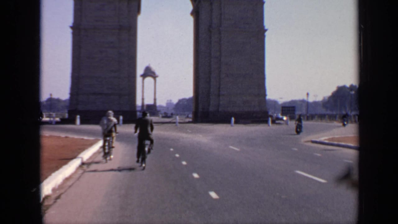 Cyclists ride away on a broad New Delhi road near India Gate, with distant pedestrians and vehicles around the landmark.