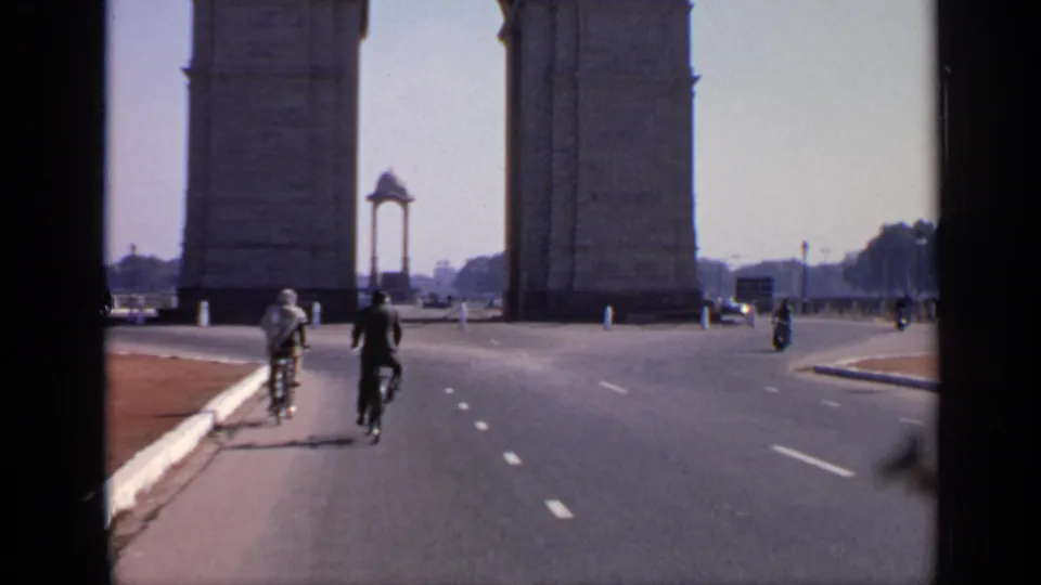 Cyclists ride away on a broad New Delhi road near India Gate, with distant pedestrians and vehicles around the landmark.