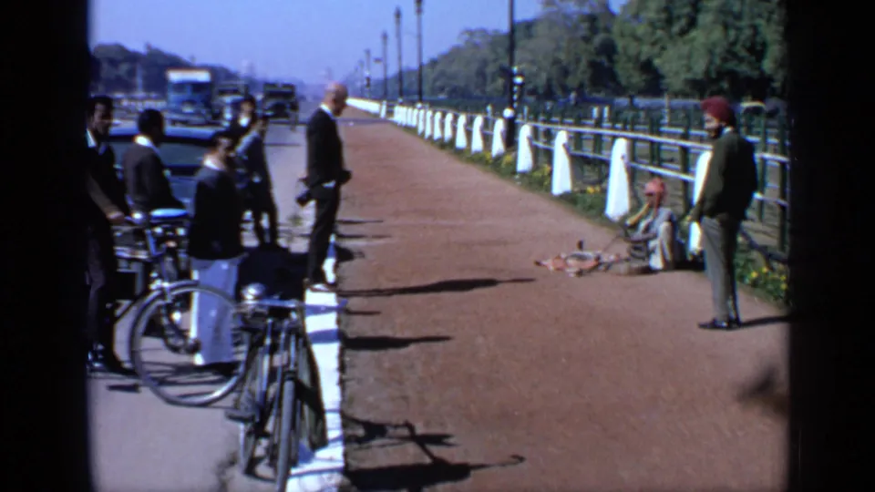 1970 India home-movie frame showing onlookers, bicycles, vehicles, and a seated performer along a public promenade.