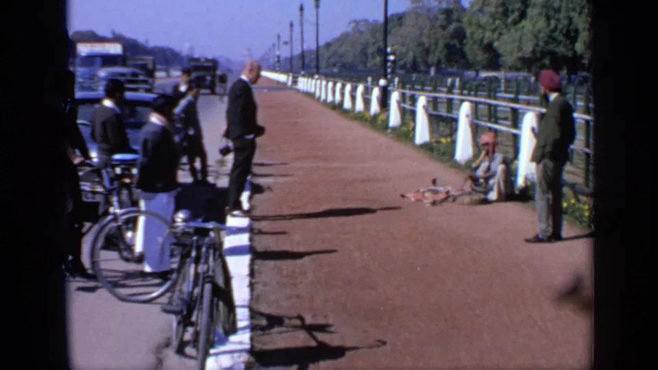 1970 India street scene with a seated musician on the pavement, people watching, bicycles at the curb, and cars along the road.