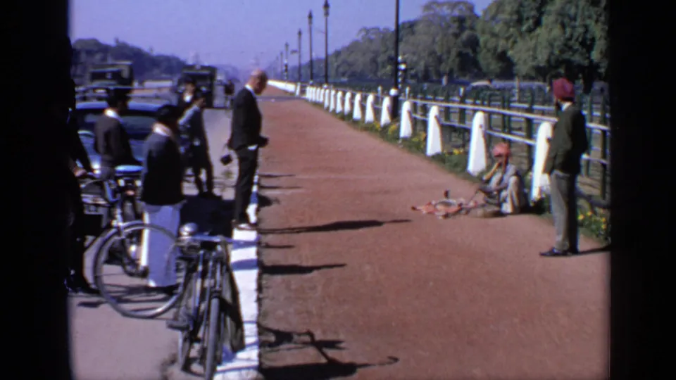 1970 India 8mm view of a seated performer with a small group watching from a sunlit walkway beside bicycles and road traffic.